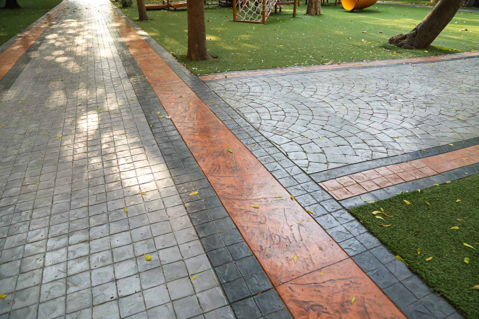 A paved walkway with gray square tiles and orange borders runs alongside a patterned pavement and artificial grass in an AZ park, featuring trees and playground equipment—similar to the durability of garage floor coating Maricopa County trusts.