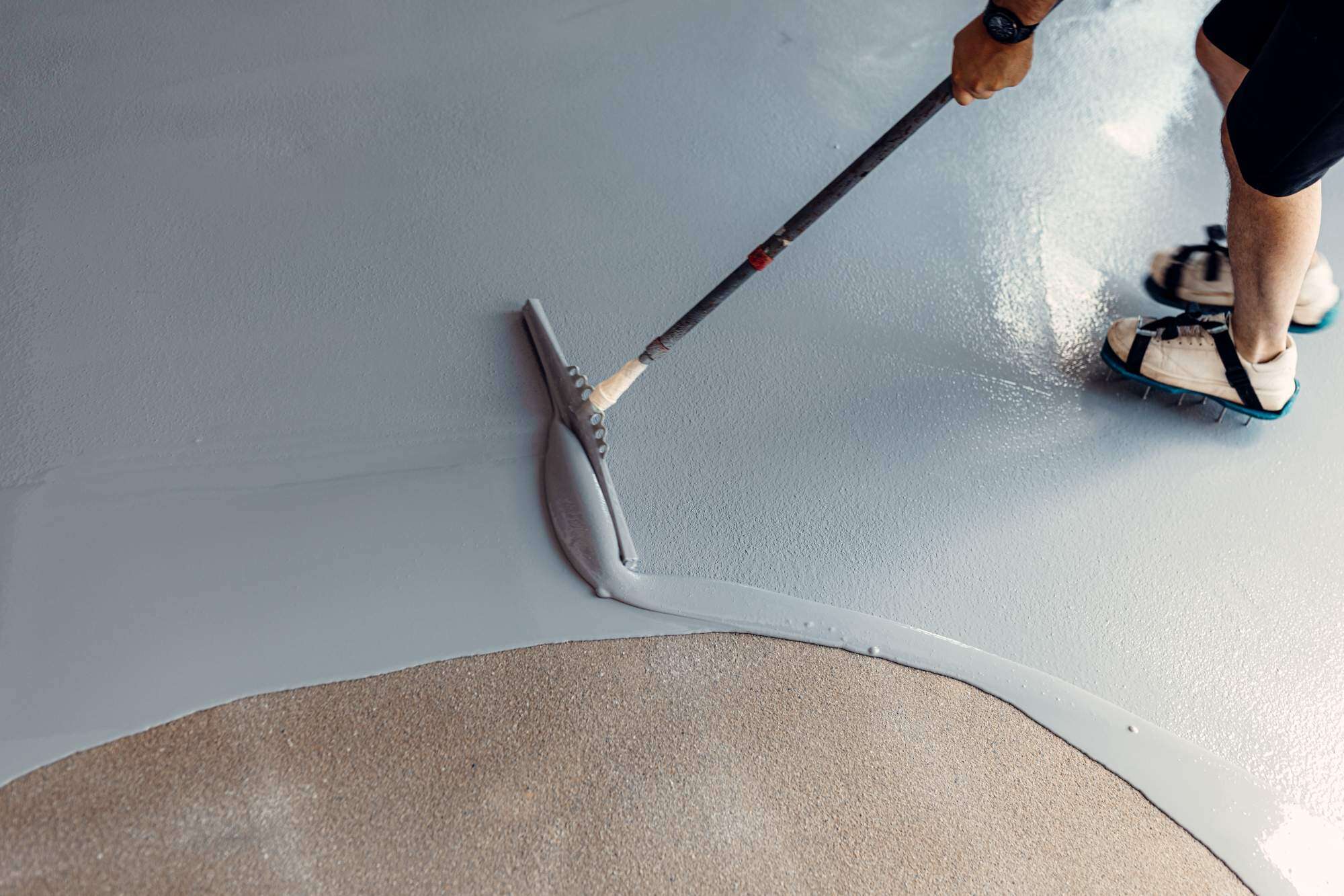 A person in sandals uses a roller on a long handle to spread gray epoxy or paint over a floor, partially covering a rough beige surface—possibly preparing for seal coating Maricopa County floors in AZ. Only their legs and footwear are visible.