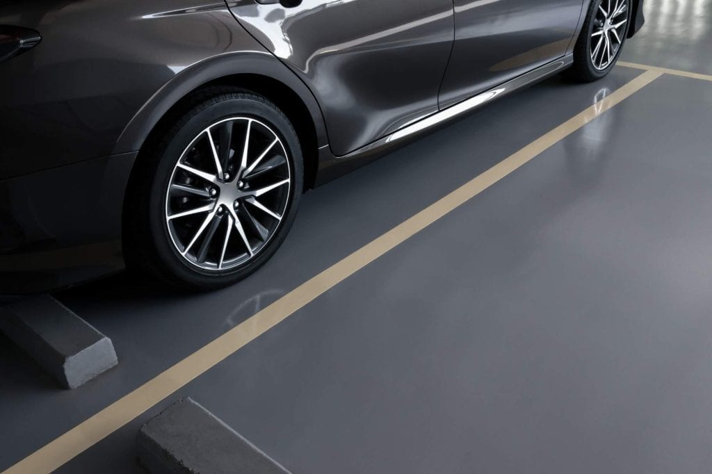 A close-up of a parked dark gray car in a parking space in AZ, showing the front wheel, part of the door, and parking lines on a smooth, seal-coated surface with wheel stops.