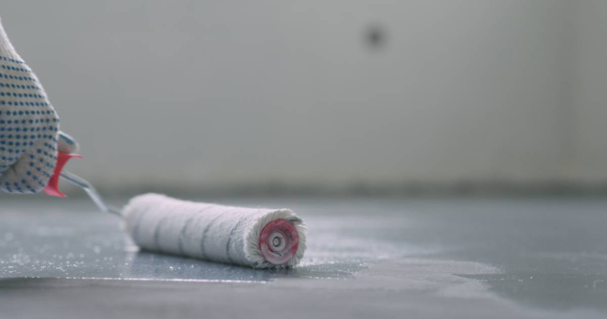 A close-up of a gloved hand holding a paint roller, spreading a light coating of garage floor coating Maricopa County on a smooth floor surface.