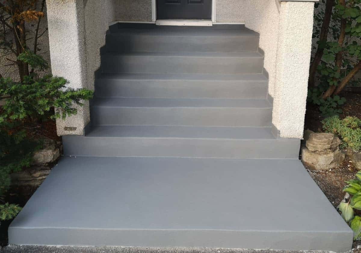 Concrete front steps and landing freshly coated in smooth gray paint, leading up to a doorway, surrounded by shrubs and plants—an excellent example of quality seal coating in Maricopa County, AZ.