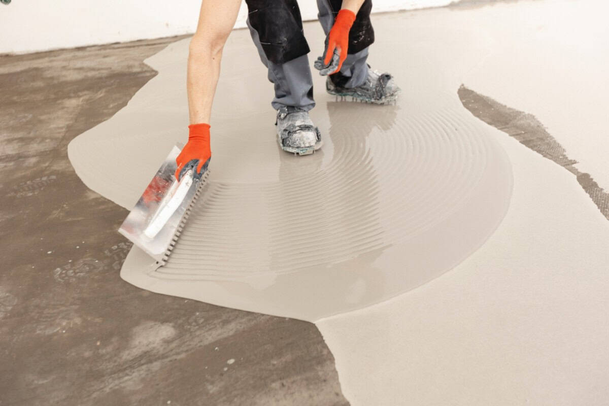 A person wearing orange gloves and work clothes spreads wet cement or floor leveling compound over a concrete surface using a notched trowel—a step often seen in seal coating Maricopa County, AZ projects.