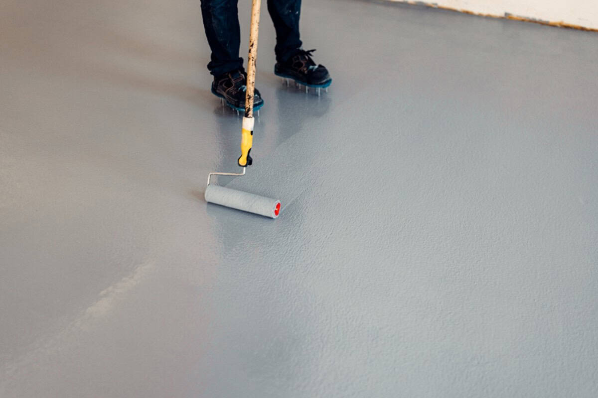 Person using a roller brush to apply a gray garage floor coating in Maricopa County, AZ; only their lower legs and shoes are visible.