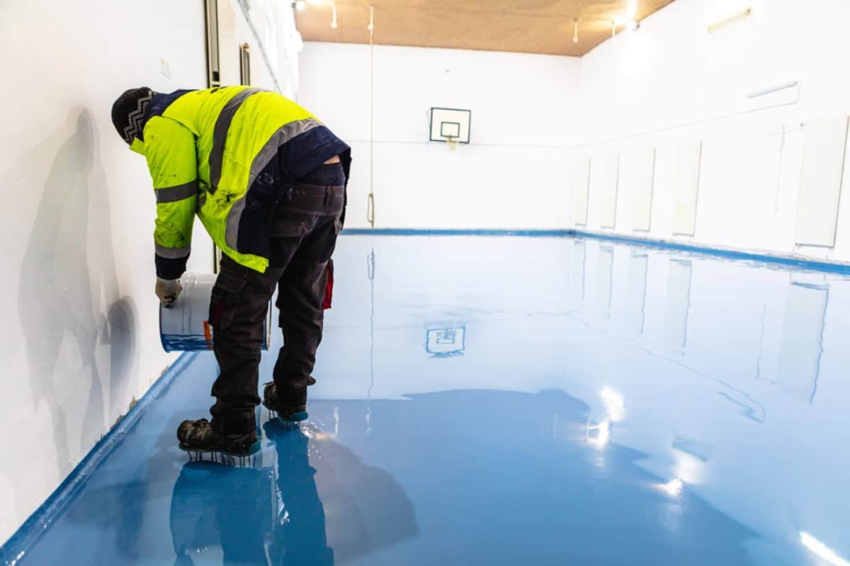 A worker in a high-visibility jacket applies seal coating Maricopa County to the edge of a shiny blue gymnasium floor using a brush, with a basketball hoop visible in the background.