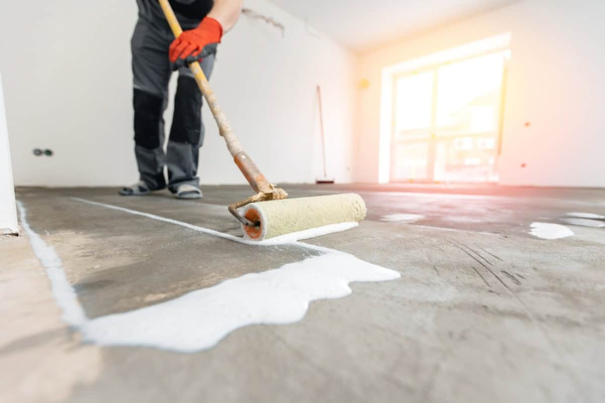 A person wearing gloves uses a paint roller to apply a white primer or sealant to a concrete floor in a bright, unfinished room, preparing it for garage floor coating in Maricopa County, AZ.
