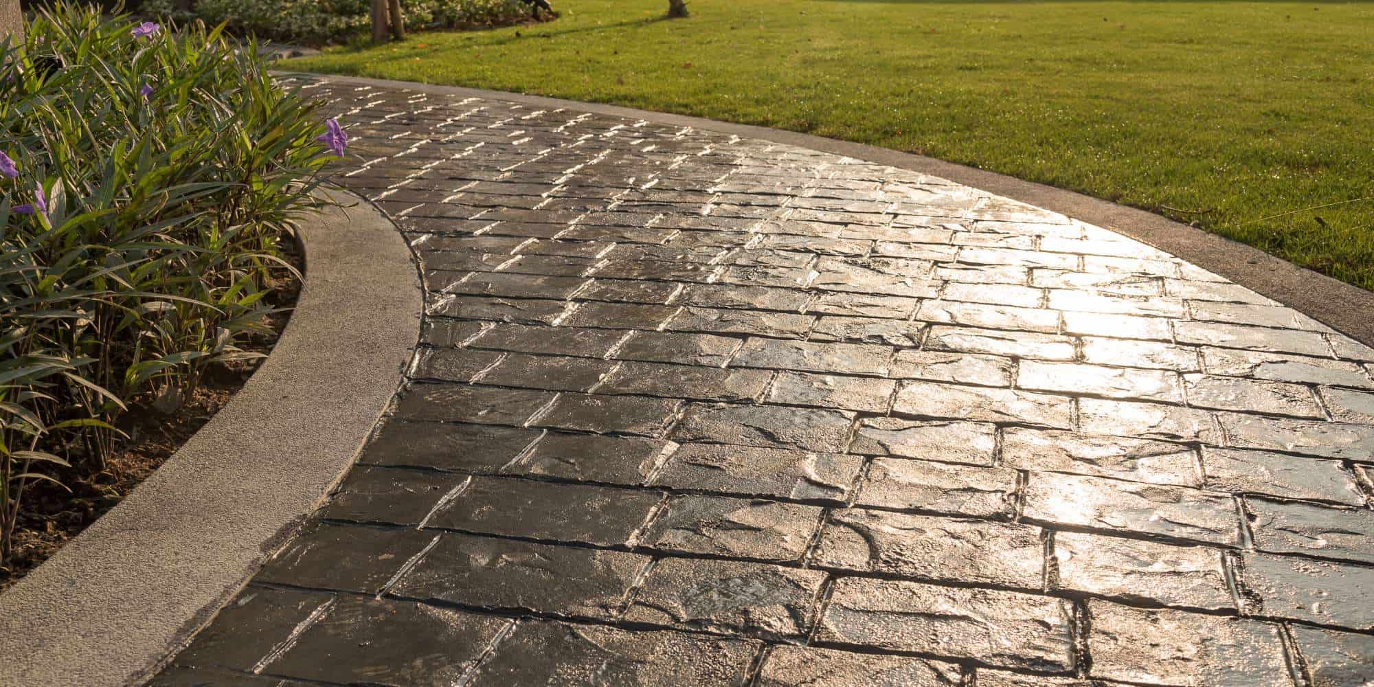 A curved, sunlit pathway made of stamped concrete runs beside a grassy lawn and a garden bed with purple flowers—showcasing expert craftsmanship like the top garage floor coating Maricopa County, AZ has to offer.
