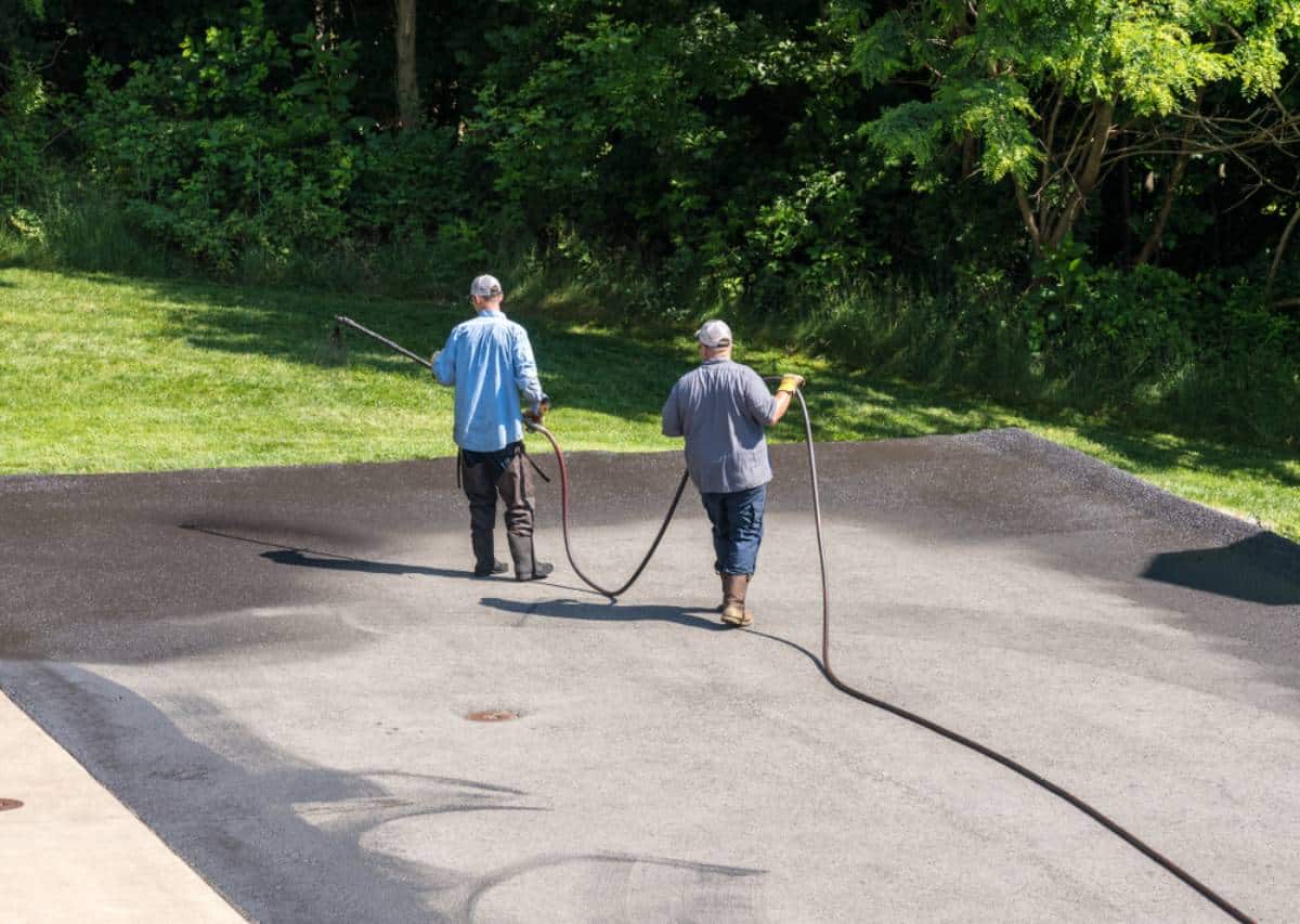 Two workers in hats and boots are spraying sealant on a driveway with long hoses, expertly applying seal coating Maricopa County, AZ. They stand on dark, freshly sealed pavement, with trees and grass in the background.