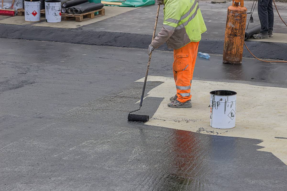 A worker in a yellow safety jacket and orange pants applies black seal coating Maricopa County material to a flat surface with a roller. Nearby are a paint bucket, gas tank, and construction materials.