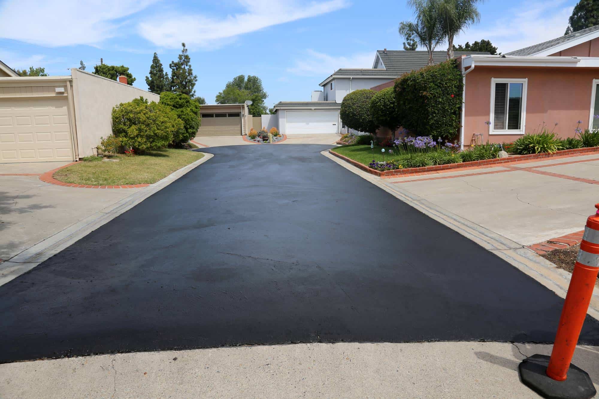 A newly paved, smooth black asphalt driveway between two homes in Maricopa County, AZ, bordered by concrete curbs and landscaping, with a bright orange traffic cone in the foreground and blue sky above—perfect for seal coating Maricopa County, AZ.