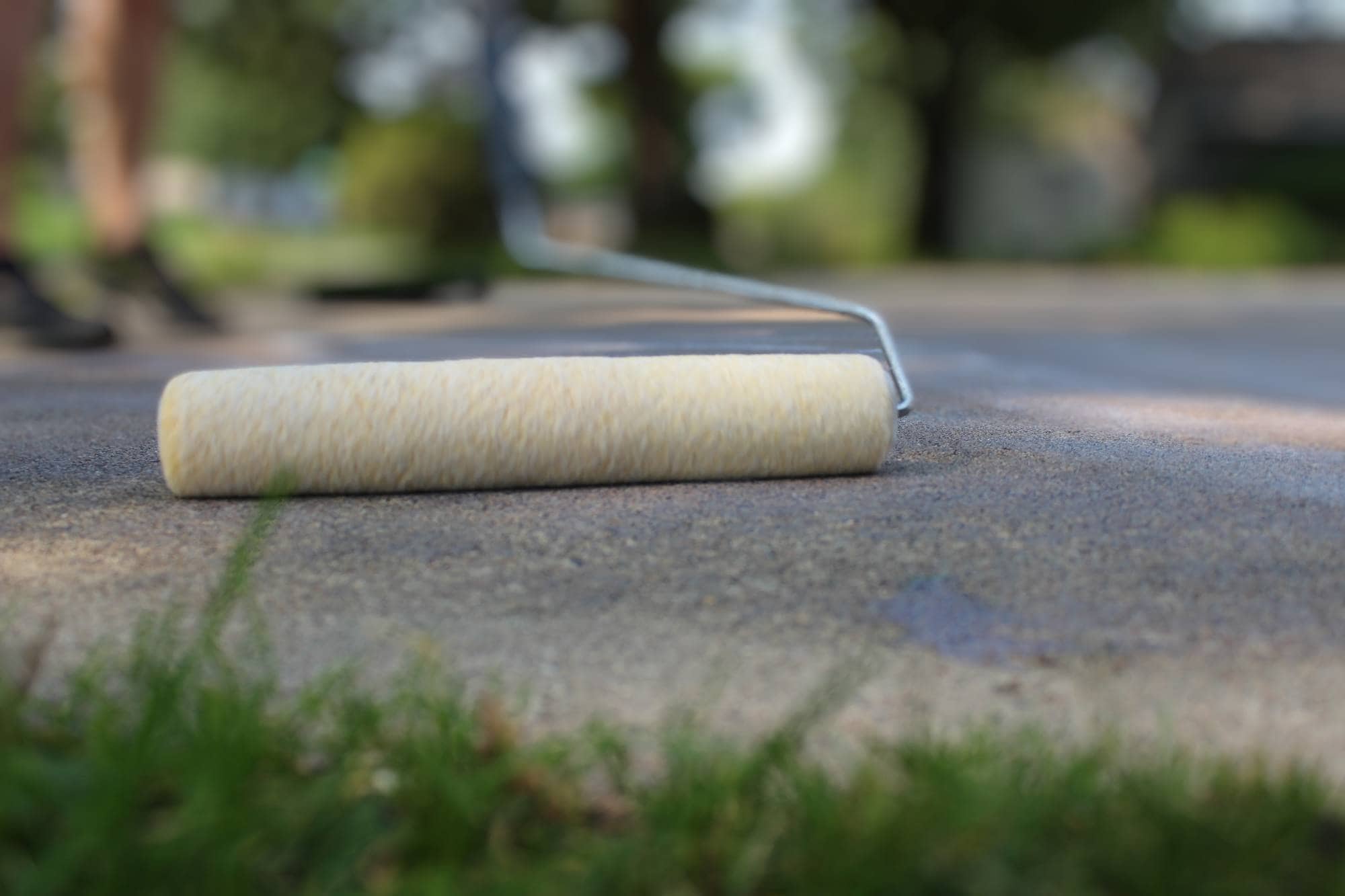 A close-up of a paint roller resting on a concrete surface outdoors in Maricopa County, with blurred grass in the foreground and trees in the background—ideal for seal coating or garage floor coating projects.
