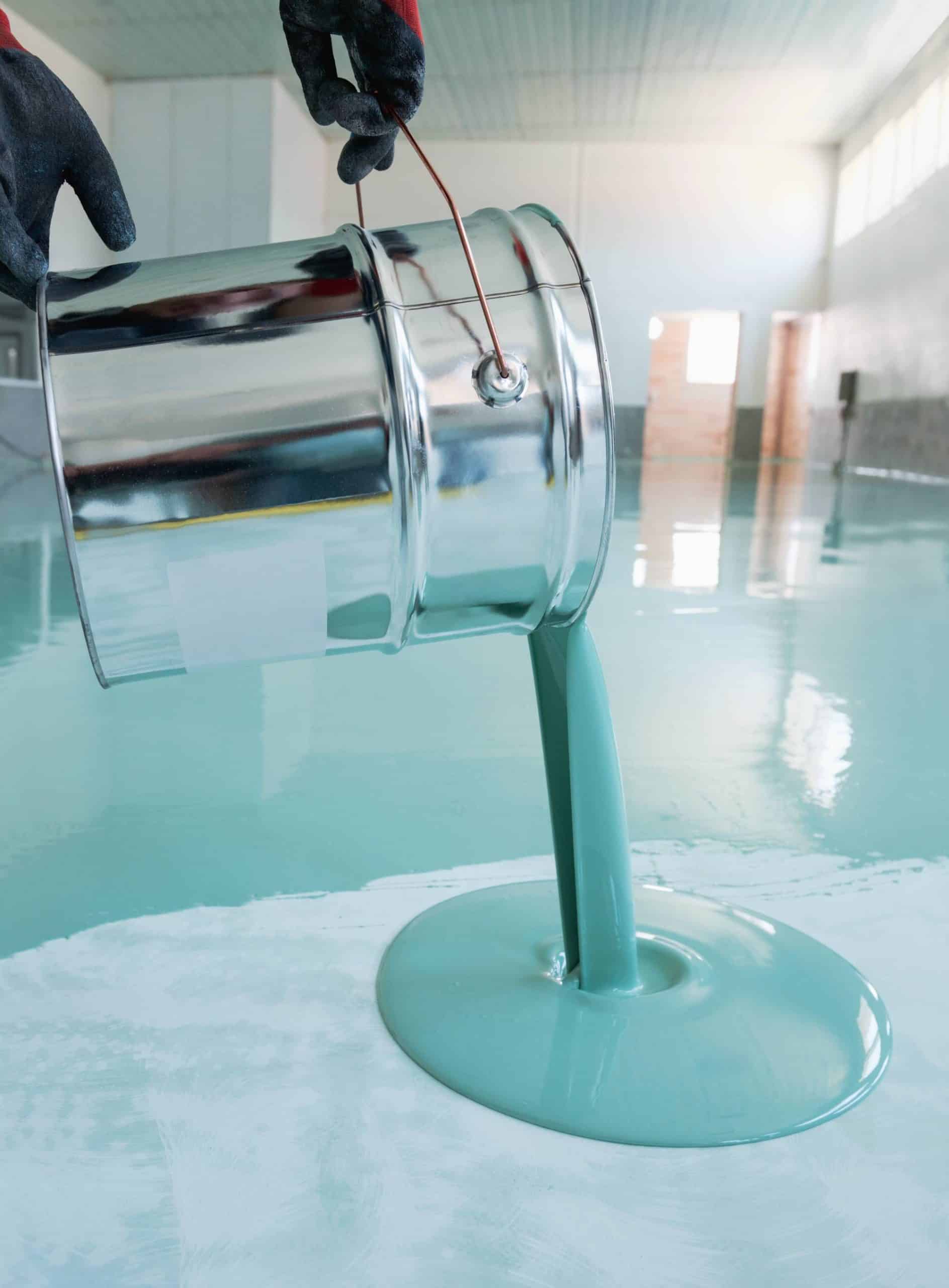 A person wearing black gloves pours a thick, light blue-green liquid from a large metal bucket onto a shiny, smooth floor in a spacious, well-lit room.