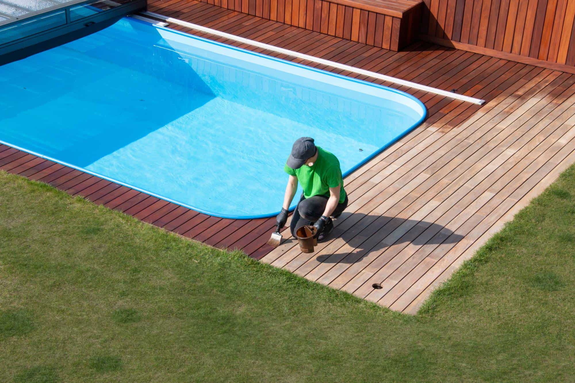 A person in a green shirt and cap is kneeling on a wooden deck next to a swimming pool, applying wood stain or sealant near the edge where the deck meets the grass.