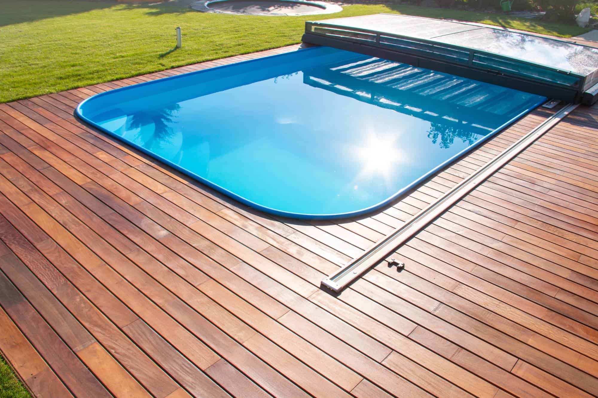 Modern backyard with a rectangular blue swimming pool surrounded by a wooden deck. The pool cover is partially retracted, and the sun reflects on the water. There is a grassy lawn in the background.